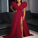 Woman wearing a flowing red maxi dress with ruffled shoulders and a high slit, posing indoors in front of a sofa.