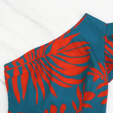 A woman smiling and posing in a teal and red tropical print maxi dress with a high slit and one-shoulder design.