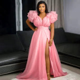 Woman wearing a flowing light pink maxi dress with ruffled shoulders and a high slit, posing indoors in front of a sofa.