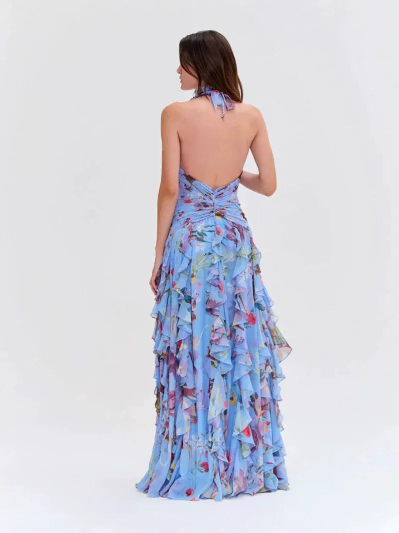 Back view of a woman wearing a light blue floral print halter neck ruffled gown with a thigh-high slit.