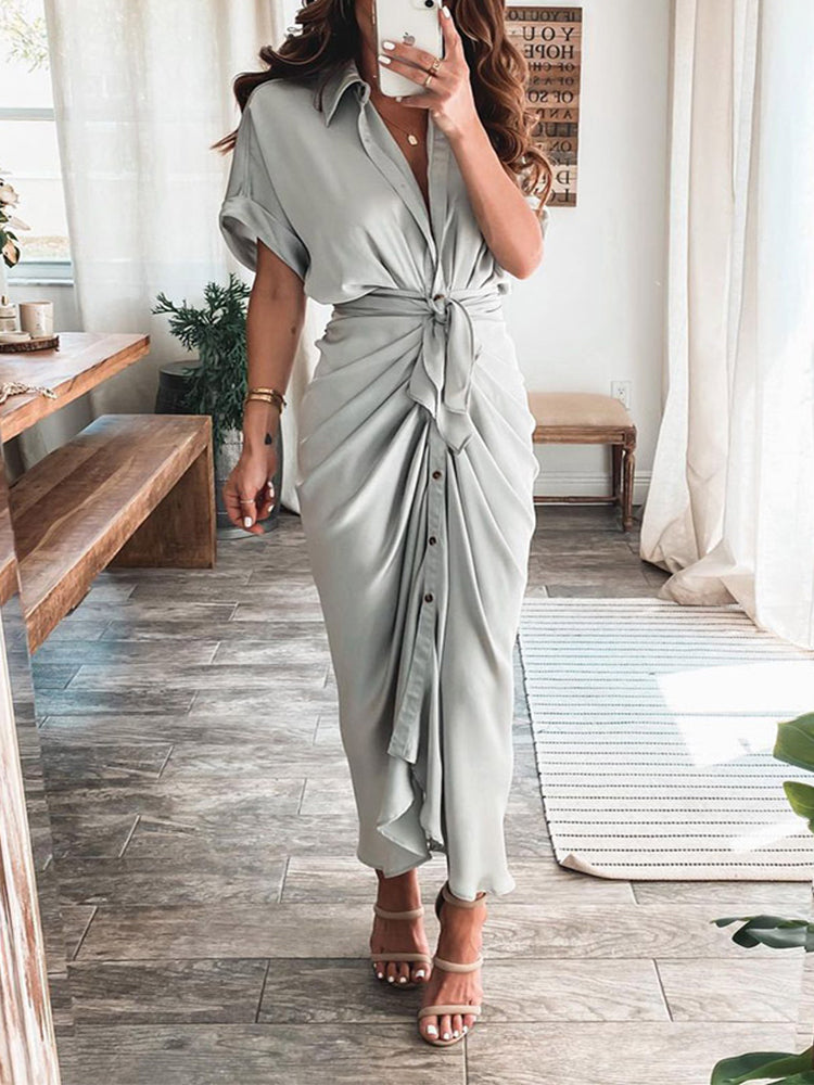 Woman wearing a light gray satin maxi dress with a collared neckline, short sleeves, a tie waist, and button-down front.
