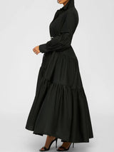 Side profile of a woman wearing a black tiered maxi dress with a collared neckline and a wide black belt.