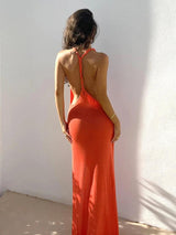 Woman from the back wearing an orange knit maxi dress with braided straps and an open back, standing against a white wall.