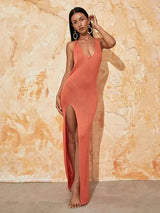 Woman wearing an orange knit maxi dress with a high slit and plunging neckline, standing against a textured wall.