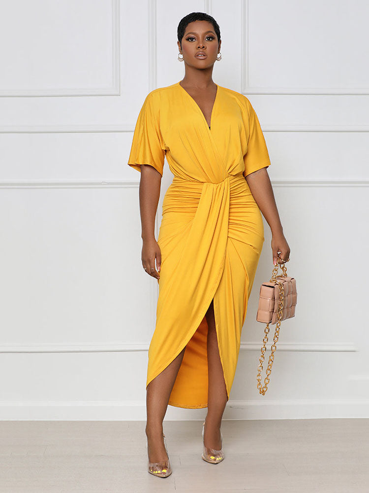 Woman wearing a yellow V-neck, short-sleeve, ruched, draped midi dress with a tulip hem, holding a beige handbag.