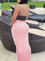 Rear view of a woman wearing a black halter crop top with a knot detail and a light pink maxi skirt.