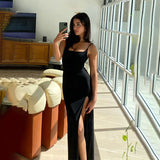 Woman wearing a black sleeveless maxi dress with a high slit, taking a mirror selfie in a sunlit room with large windows and modern furniture.