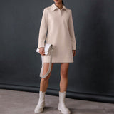 Full body shot of a woman wearing a light beige long-sleeved collared shift dress, white handbag, and white ankle boots.