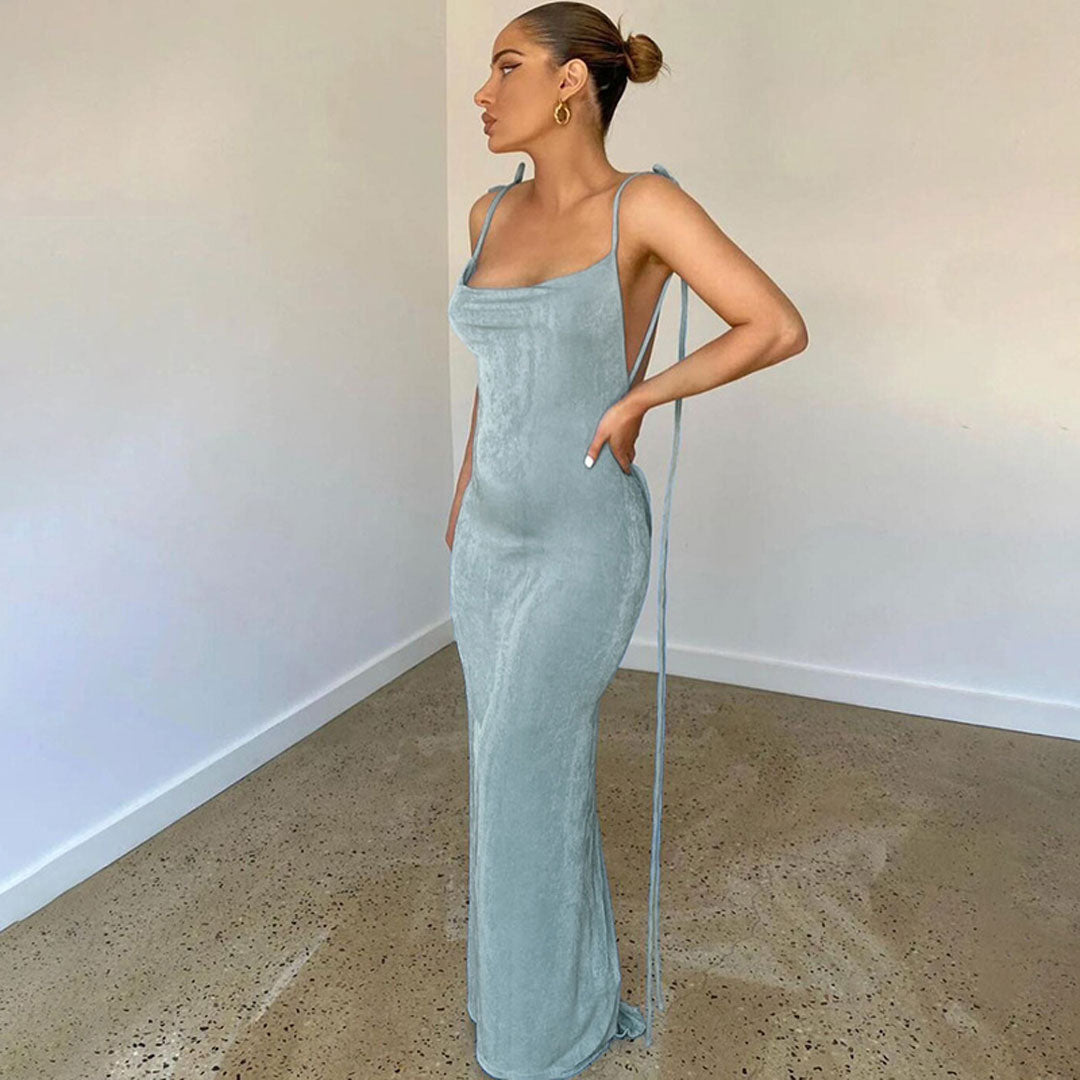 Woman in a light blue sleeveless maxi dress with a cowl neckline and backless design, standing in a room.