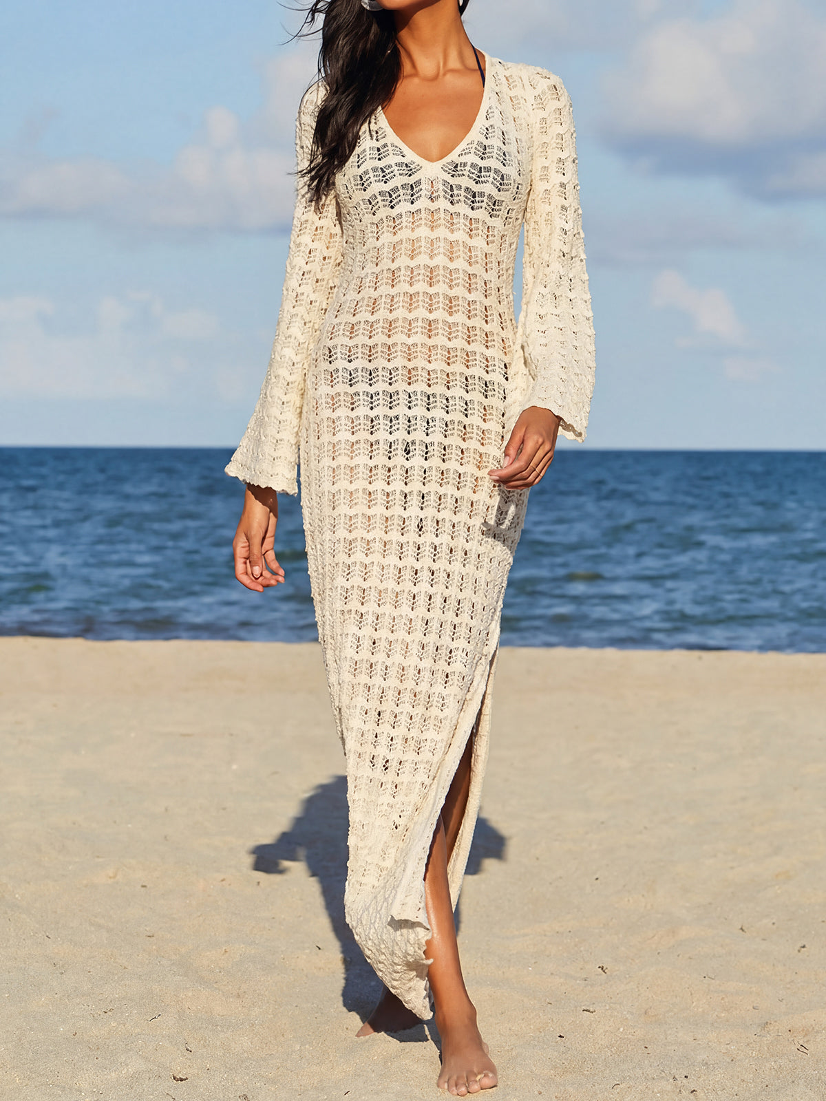 Woman wearing a long-sleeved cream crochet knit maxi dress with a V-neck and side slit, standing on a beach.