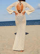 Back view of a woman wearing a long-sleeved cream crochet knit maxi dress with an open back detail and bell sleeves, standing on a beach.