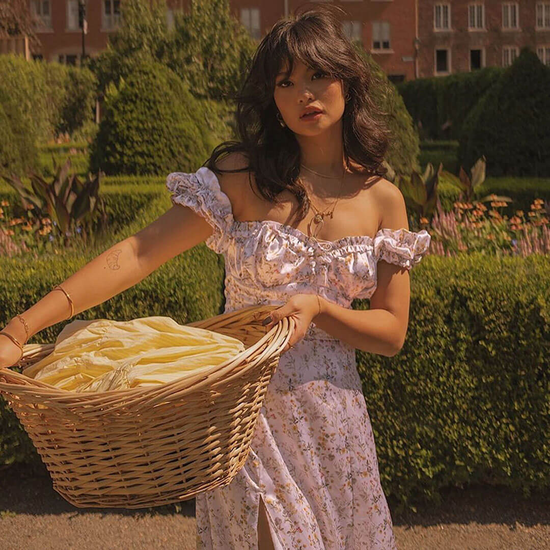 Woman in a white floral off-the-shoulder maxi dress with a high slit holding a basket of laundry in a garden.