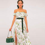 Woman in a white floral off-the-shoulder maxi dress with a high slit and black belt, holding a green handbag.