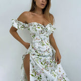 Woman in a white floral off-the-shoulder maxi dress with a high slit, standing against a white wall.