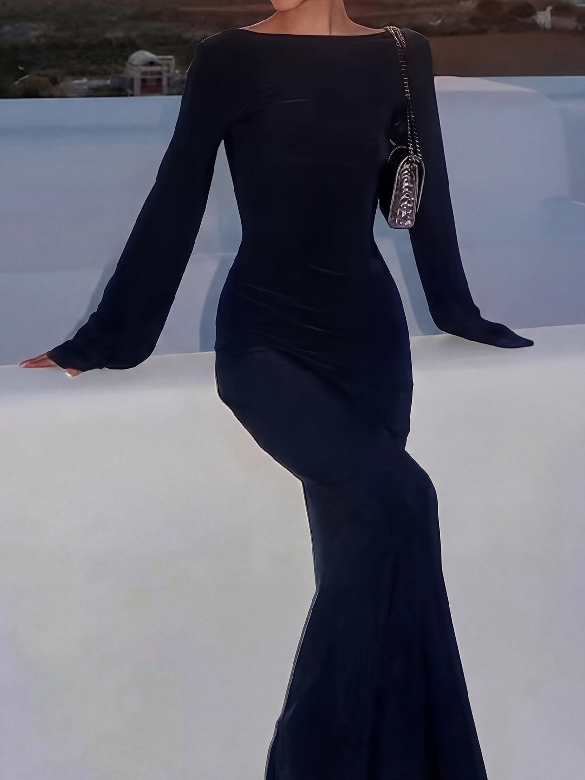 Woman in a black long-sleeved maxi dress with flared sleeves, leaning on a white ledge with a small handbag.