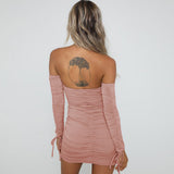 Back view of a woman wearing a pink off-the-shoulder ruched mini dress with long sleeves and a tie detail at the bust.