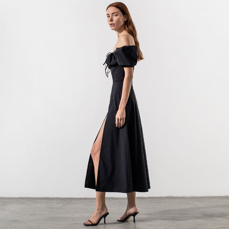 Woman wearing a black off-the-shoulder midi dress with puffed sleeves and a high slit, side view.