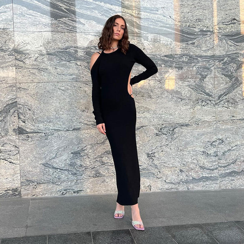 Woman in a long-sleeved black maxi dress with a cut-out shoulder detail, standing against a marbled wall.