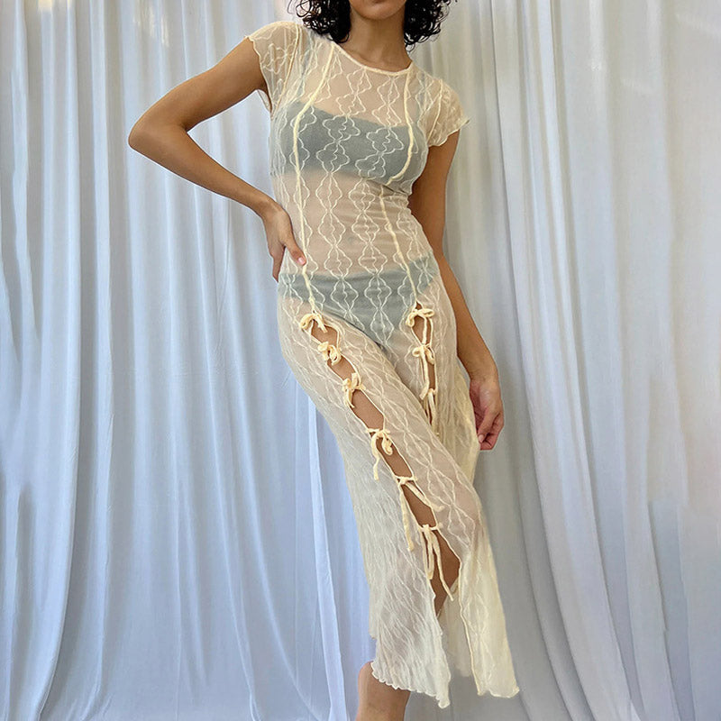 Woman wearing a sheer cream midi dress with short sleeves and tie closures on the side, standing in front of a white curtain.