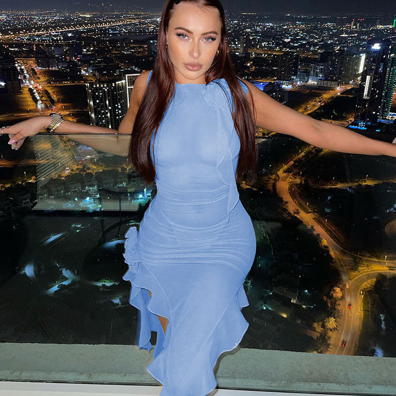 Woman wearing a light blue asymmetrical midi dress with ruffle detailing and ruching, standing on a balcony at night with a city skyline in the background.