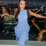 Woman wearing a light blue asymmetrical midi dress with ruffle detailing and ruching, standing on a balcony at night with a city skyline in the background.