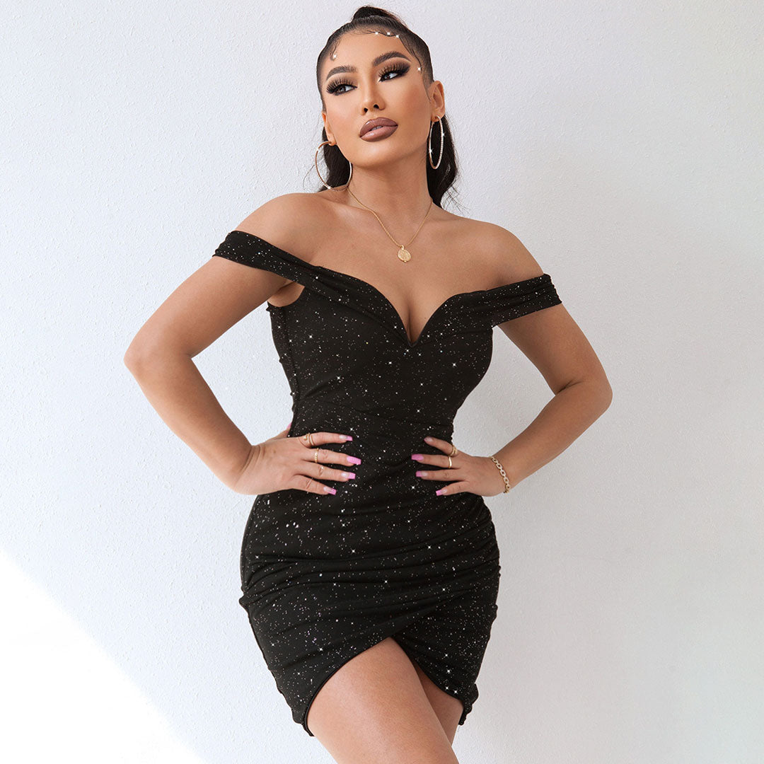 Front view of a woman wearing a black off-the-shoulder sparkly mini dress with ruched detailing and an asymmetrical hemline, with one hand on her chest.