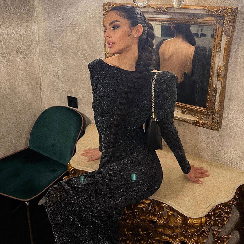 Woman in a black sparkly long-sleeved dress with an open back, posing by a mirror.