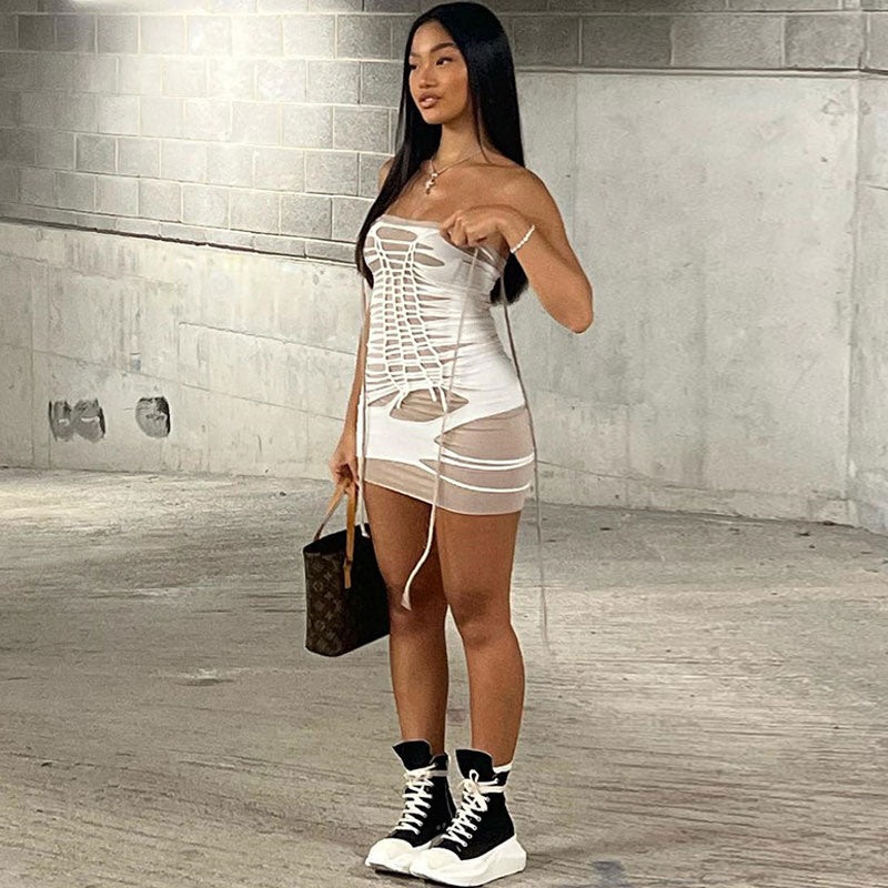 Side view of a woman in a white strapless bodycon mini dress with cut-out details and a lace-up front, holding a brown designer handbag.