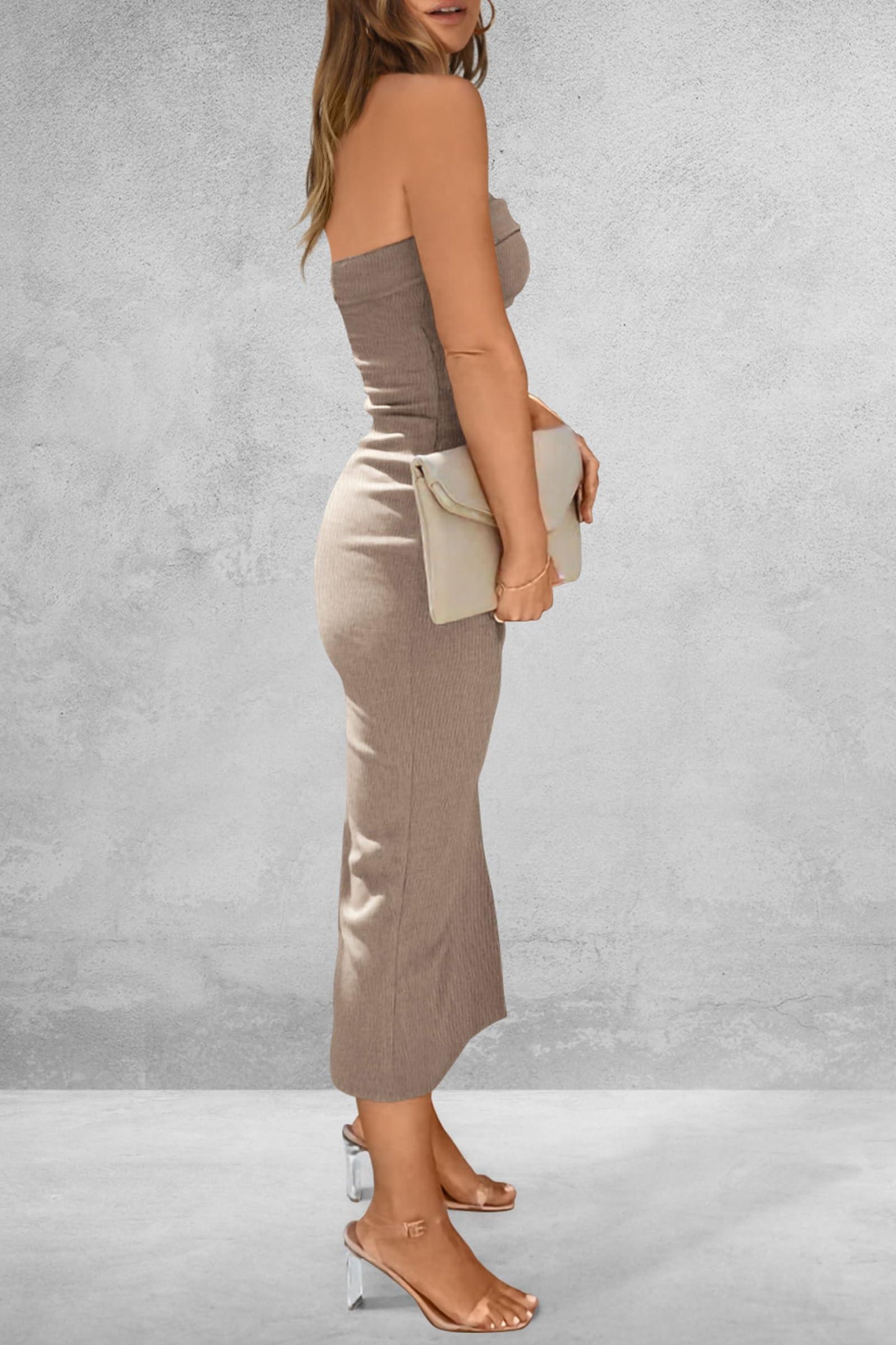 Side view of a woman wearing a taupe strapless ribbed knit maxi dress with a side slit, holding a cream clutch and wearing clear strap heels.