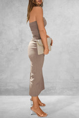Side view of a woman wearing a taupe strapless ribbed knit maxi dress with a side slit, holding a cream clutch and wearing clear strap heels.