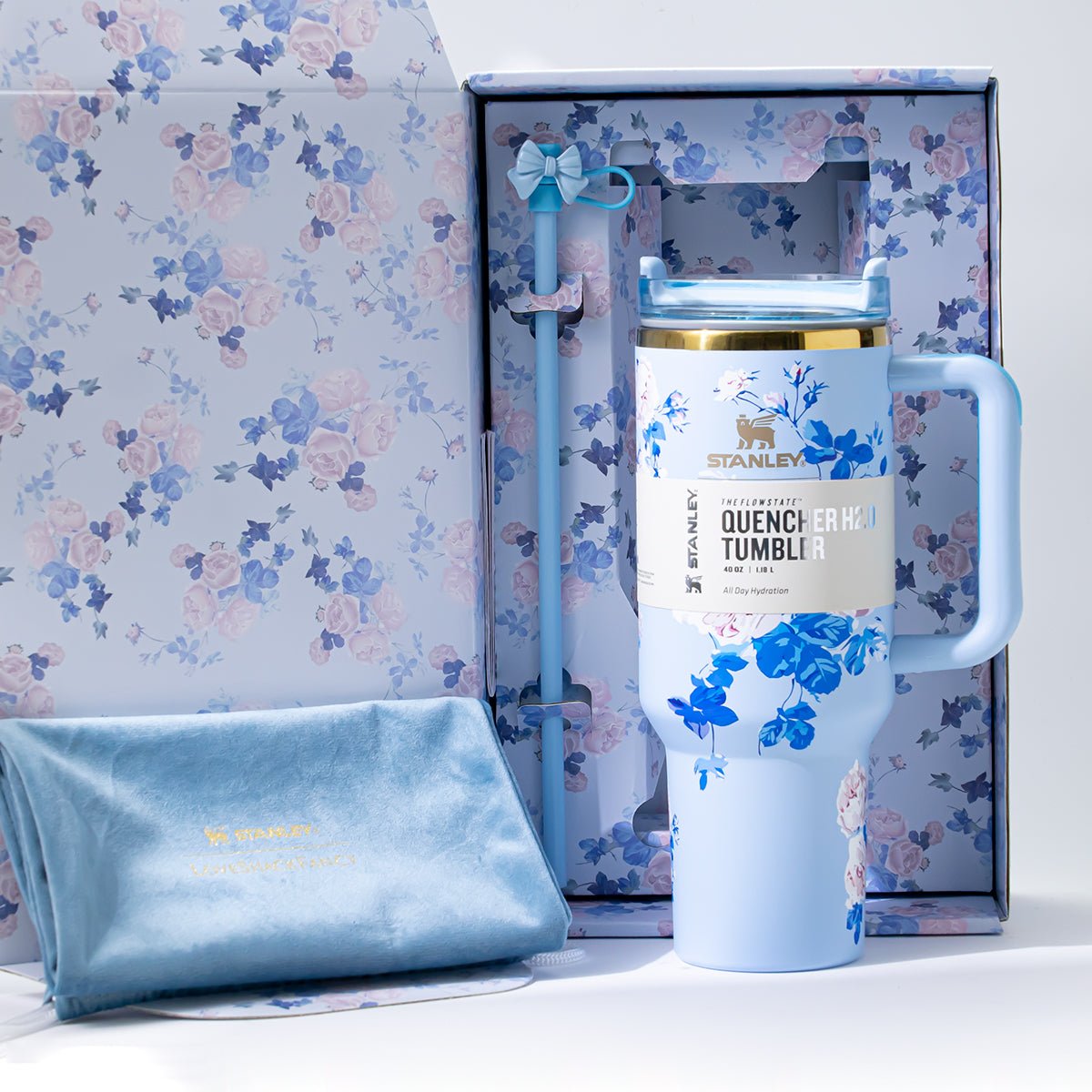 An open floral-patterned gift box containing a light blue Stanley Quencher H2.0 Tumbler, a straw, and a soft blue cloth pouch.