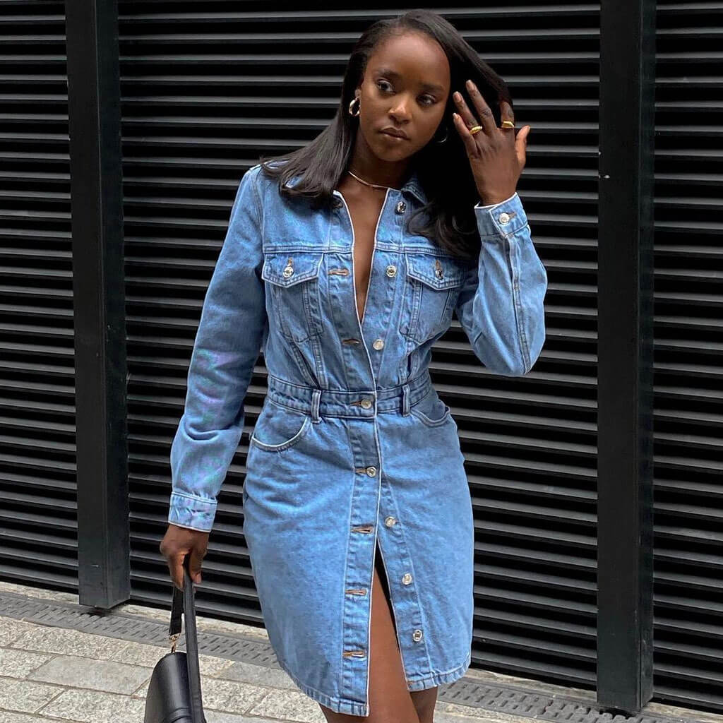 Woman in a denim shirt dress with a belted waist, standing against a black slatted background.