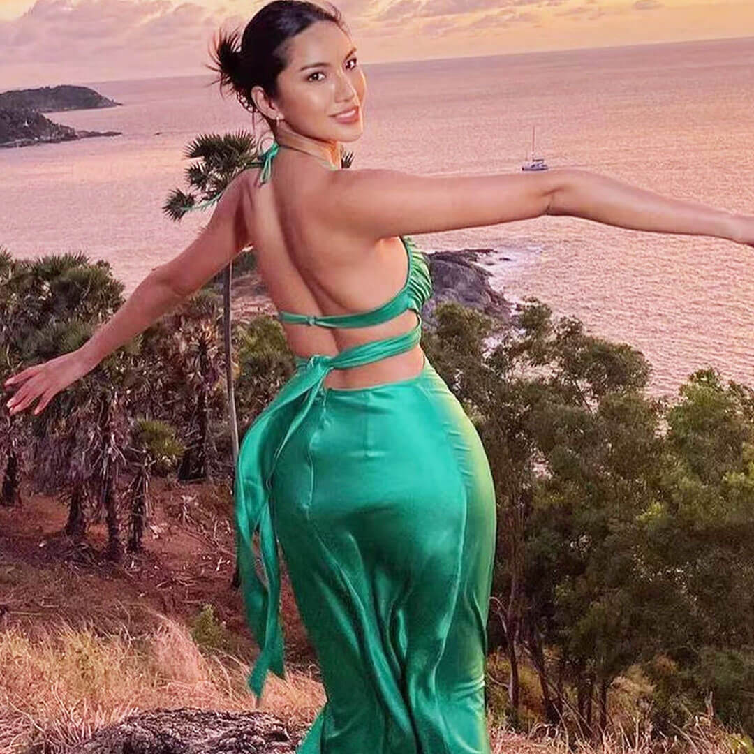 Back view of a woman in a green satin maxi dress with a halter neckline and tie-back closure, with arms outstretched.