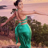 Back view of a woman in a green satin maxi dress with a halter neckline and tie-back closure, with arms outstretched.