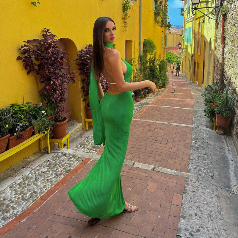 Woman wearing a green one-shoulder velvet maxi dress with a high slit and open back, posing on a cobblestone street.