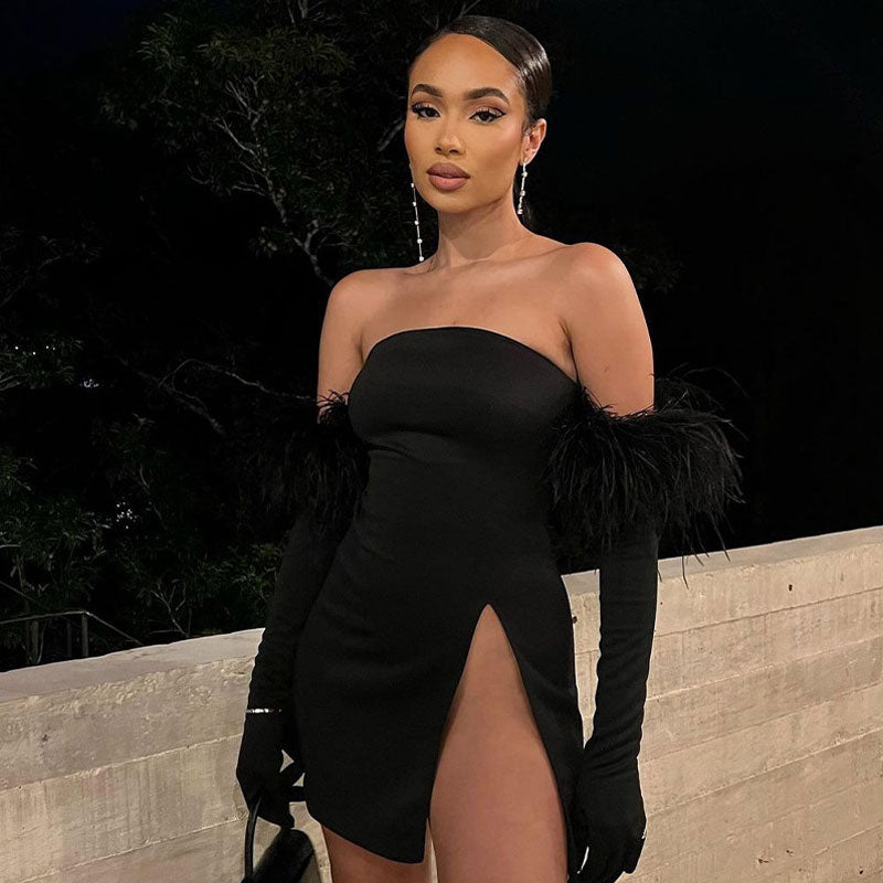 Woman wearing a black strapless mini dress with a high slit and long feather-trimmed gloves, posing outdoors at night.