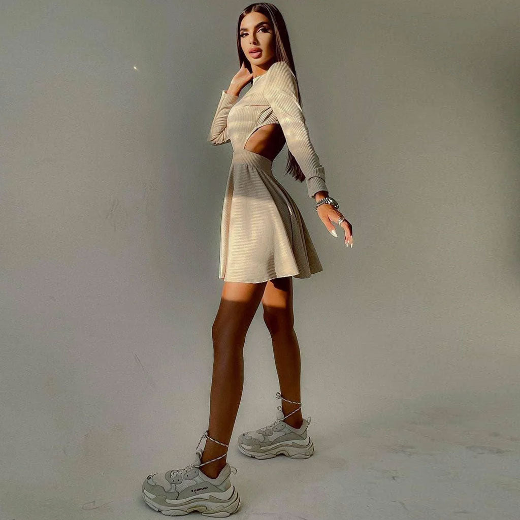 Woman wearing a long-sleeved beige ribbed knit fit and flare dress with a crew neck and cut-out detail, posing with one leg extended.