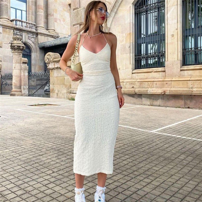 Woman wearing a white textured midi dress with a halter neckline and a side slit, paired with white sneakers and a cream handbag.