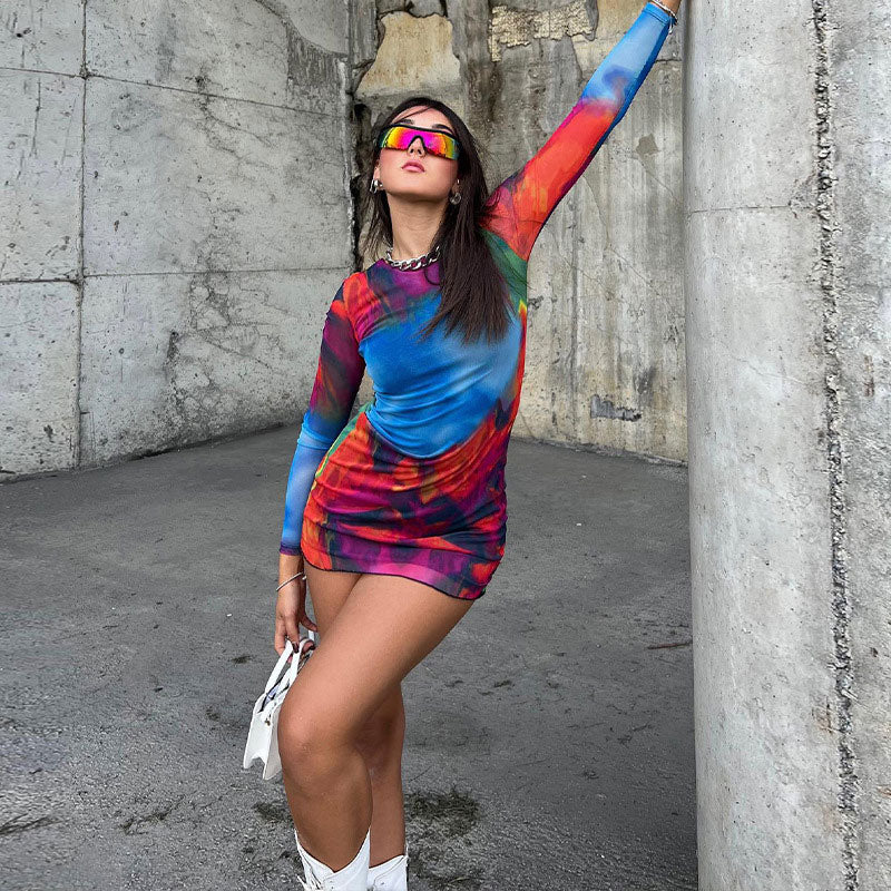 Woman wearing a colorful tie-dye mesh mini dress with long sleeves and sunglasses, leaning against a concrete pillar.