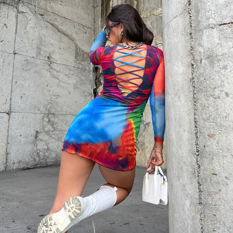 Woman wearing a colorful tie-dye mesh mini dress with long sleeves and an open criss-cross back, leaning against a concrete pillar.