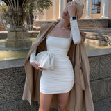Woman in a white long-sleeved ruched mini dress with a tan trench coat and a white clutch, standing next to a fountain.