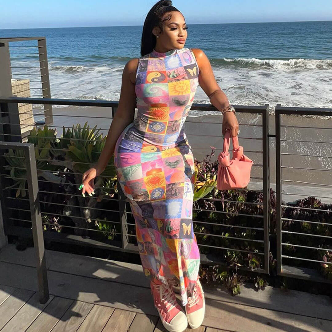 Woman wearing a colorful patchwork print maxi dress with a mock neck and sleeveless design, standing on a balcony overlooking the ocean.