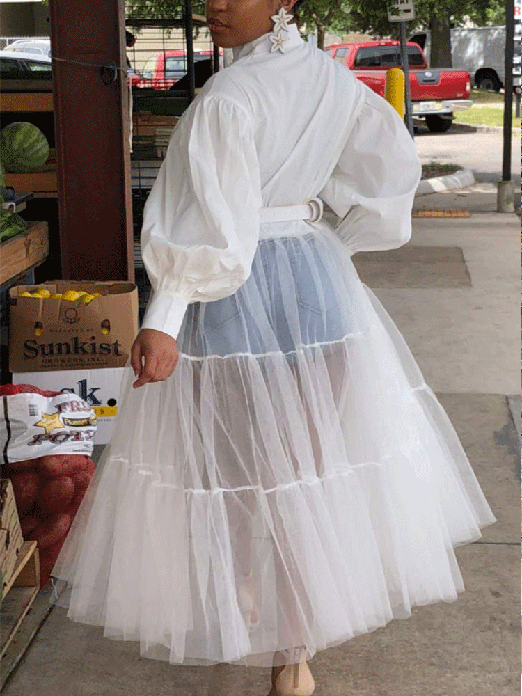 Rear view of a woman wearing a white shirt dress with a sheer white tiered tulle skirt overlay and denim shorts.