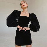 Woman wearing a black velvet mini dress with puff sleeves and a square neckline, posing with hands on hips.