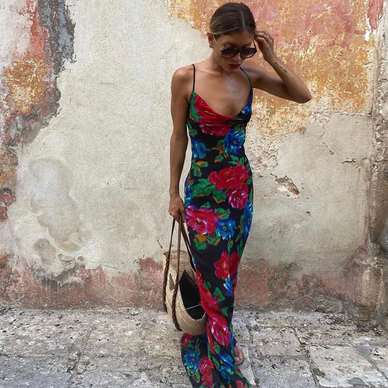 Woman wearing a black floral print maxi slip dress with a cowl neckline and spaghetti straps, holding a woven tote bag.