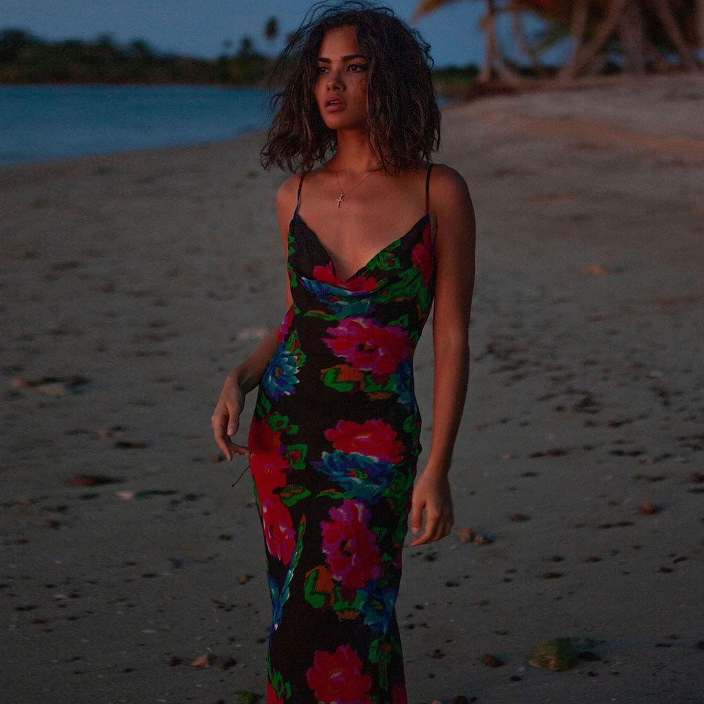 Woman wearing a black floral print maxi slip dress with a cowl neckline on a beach at dusk.