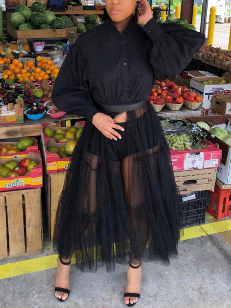 Woman wearing a black button-down shirt dress with a sheer black tiered tulle skirt overlay and black bottoms, standing in front of fruit stands.