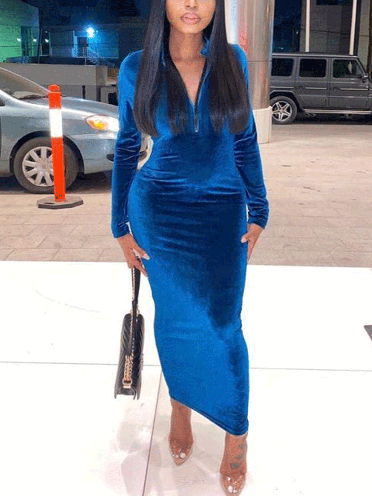 A woman stands outdoors wearing a royal blue velvet midi dress with a quarter-zip neckline and carries a black handbag.