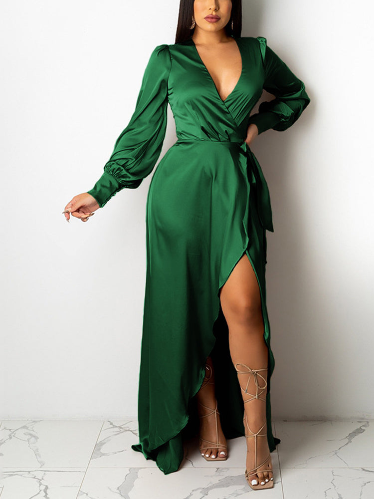 Woman wearing a forest green satin wrap maxi dress with a high slit and long puff sleeves, posing against a white wall.